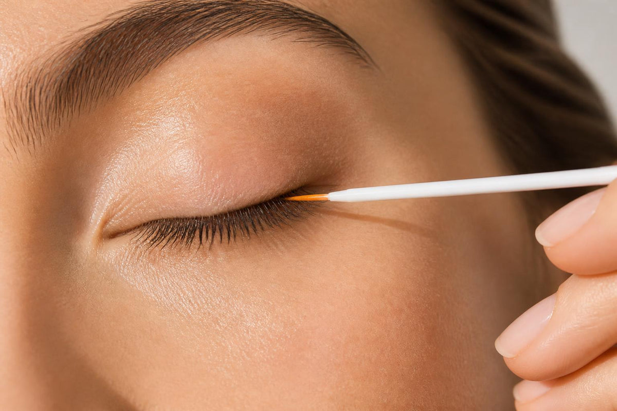 Fine-tip LashOriginals applicator making one light stroke along a closed upper lash line.
